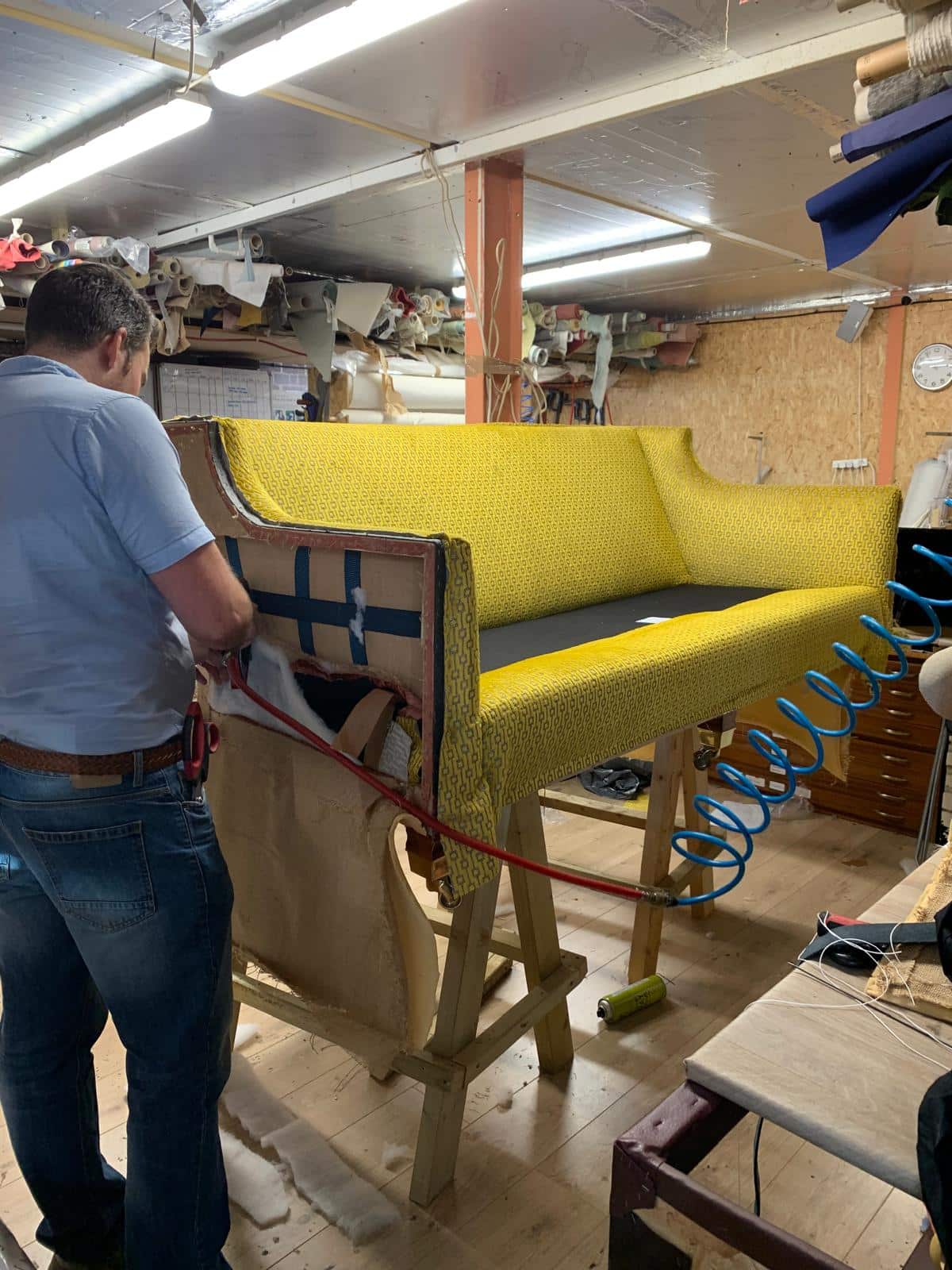 Upholsterer fitting yellow patterned fabric to a sofa with exposed side framing and webbing in a workshop.