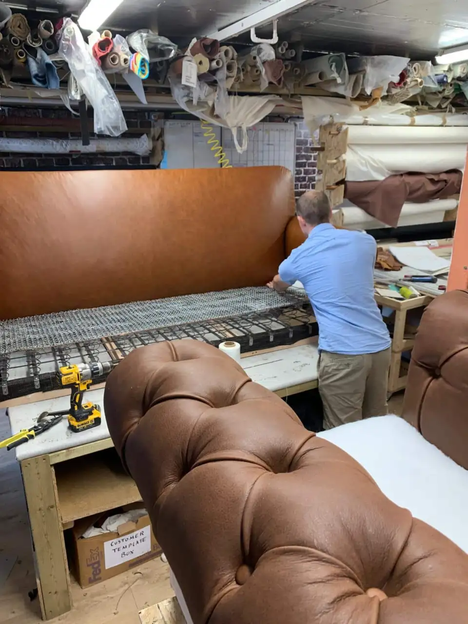 Upholsterer working on an exposed sofa seat base, with brown buttoned upholstery in the foreground.